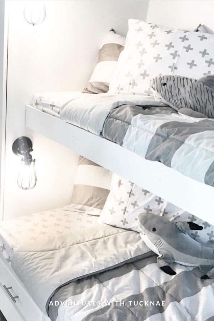 Neutral-toned RV bunk beds with soft gray and white patterned bedding, including crosses and stripes. A plush shark toy adds a fun touch, and exposed bulb wall sconces provide warm, focused light on each bunk.