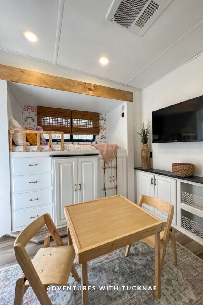A multifunctional RV kids’ room with a light wood play table and chairs in the foreground. In the back, there’s a high loft bed framed by rainbow wallpaper, built-in drawers and cabinets underneath, and a decorative rope ladder for access.