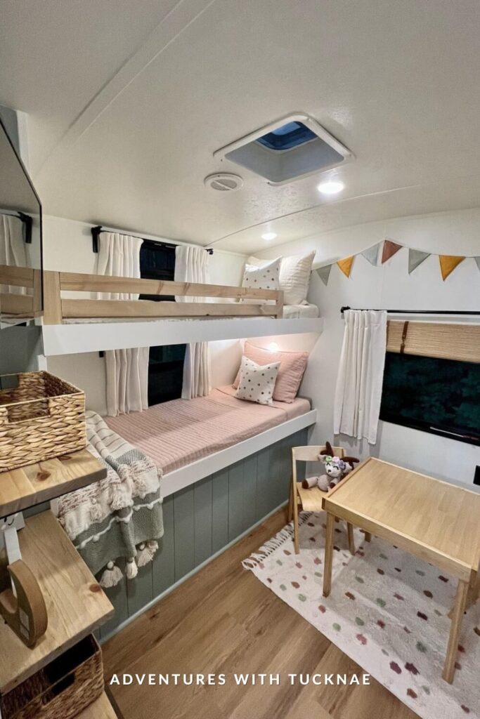 Modern RV bunk space with soft pink bedding and a small wooden table and chairs set on a speckled rug. The bunks are framed with natural wood safety rails and flanked by white curtains and triangular bunting in muted colors.