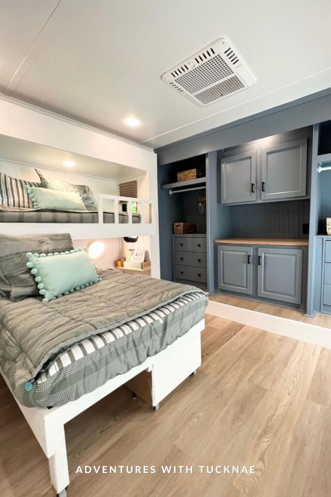 A crisp, coastal-inspired RV bedroom with gray built-in cabinetry and open shelving across from a set of wide bunk beds. The lower bunk is extra wide and dressed with striped bedding and a blue pom-pom accent pillow.