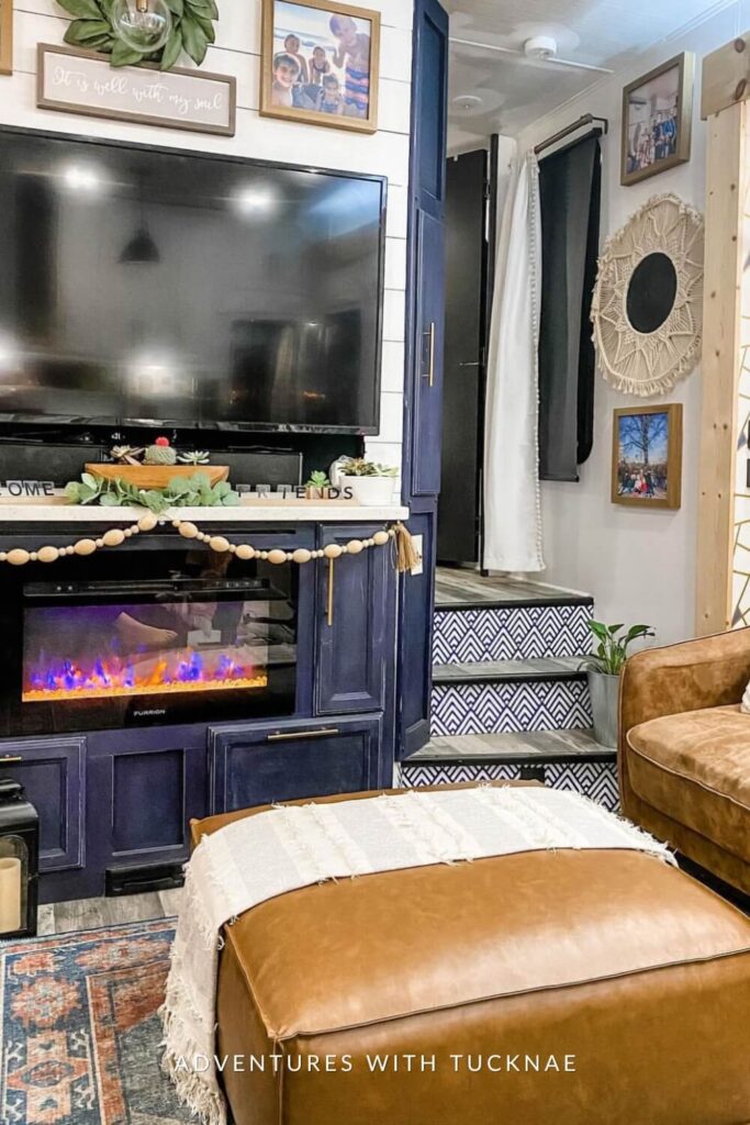 A stylish RV living area with a black entertainment center and electric fireplace below a 'Welcome Friends' sign, adjacent to a cozy sofa corner with a macrame wall hanging.