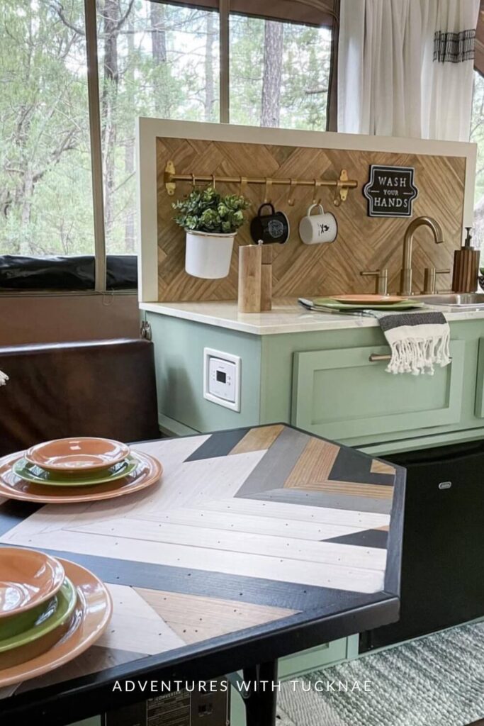 A fresh and natural RV dining nook with mint green cabinetry and a geometric backsplash, featuring a wood dining table set with orange plates and a window view of the forest.