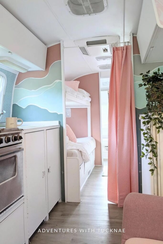 A serene RV sleeping area with ocean wave wall art above a window, a pink bunk bed alcove, and a calming color palette, complete with hanging plants and natural light.