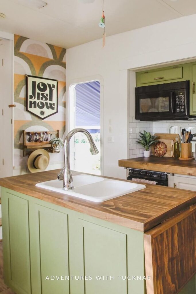 A charming RV kitchen island with a farmhouse sink, green cabinets, and a wooden countertop, set against a backdrop of a stainless steel refrigerator and white cabinetry.