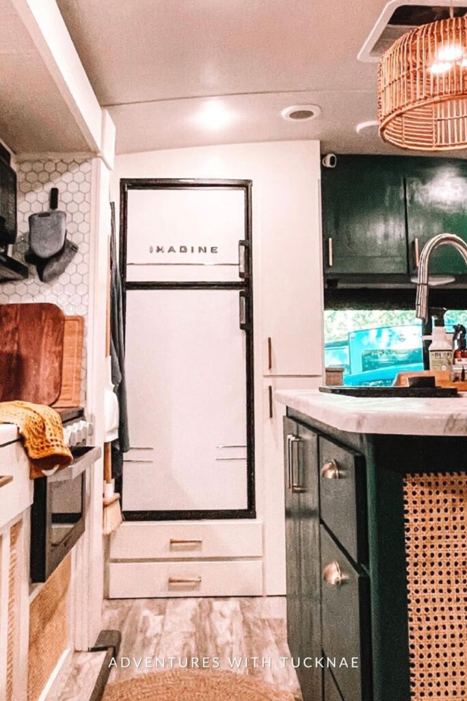 A chic RV kitchen corner with hexagonal tile backsplash, a white fridge, and green upper cabinets, illuminated by a unique wicker lampshade.