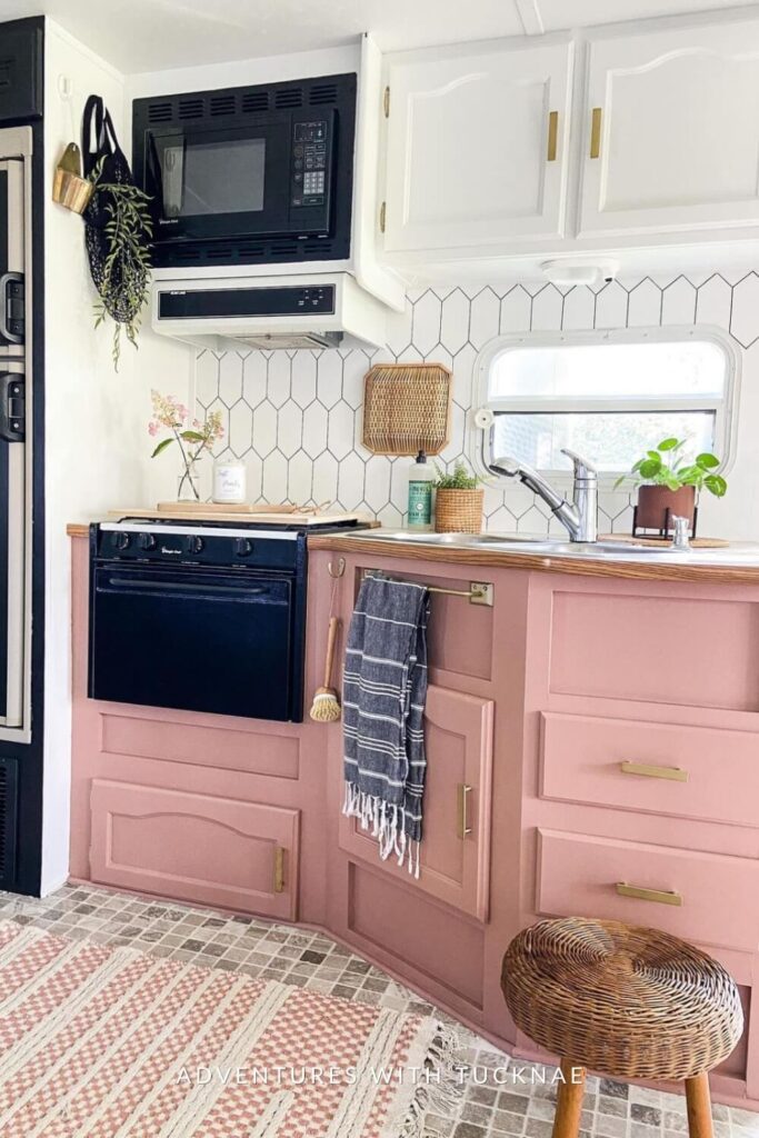 A chic RV kitchen featuring a black stove and oven, with pink cabinets and a white hexagonal tile backsplash, complemented by wicker lampshade and a sink with a potted plant.