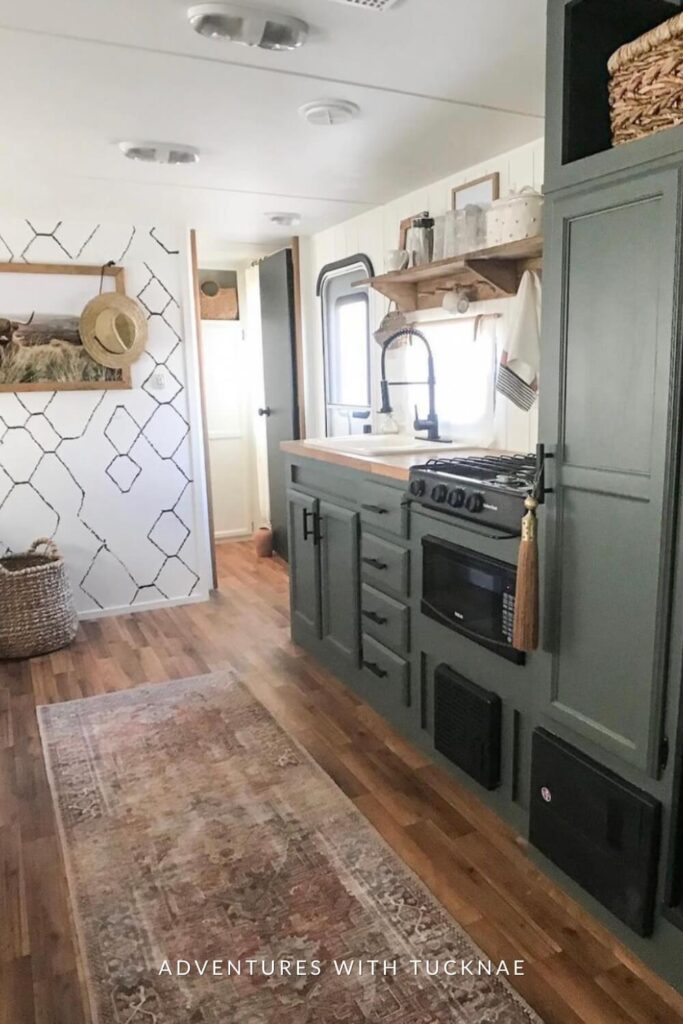 Rustic and cozy RV interior with a warm leather sofa, a white drop-leaf table under a window with a hexagonal pattern accent wall, and a kitchen area featuring green cabinetry and modern appliances.