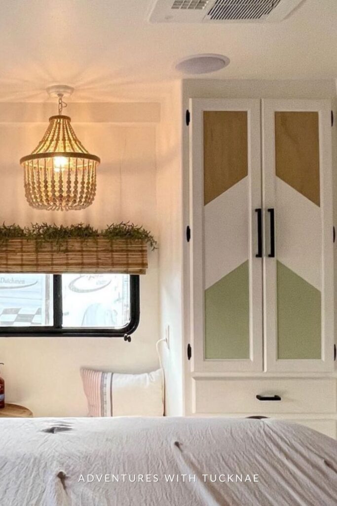 Elegant RV bedroom with a chandelier, white custom cabinets featuring geometric designs, a window with a hanging greenery planter, and a cozy bed with white linens.