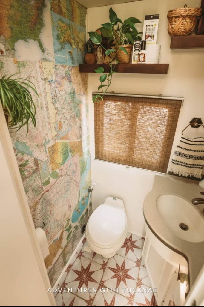 Compact RV bathroom with a map-themed shower curtain, bamboo window shade, white sink, and toilet, enhanced by a decorative striped towel and a potted plant.