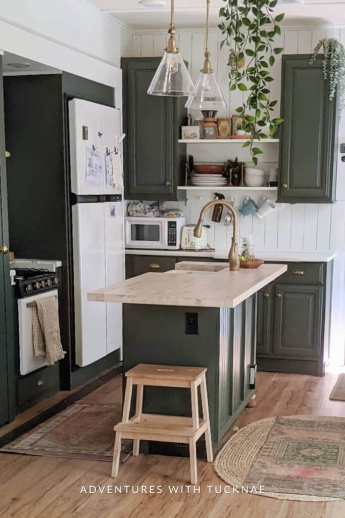 A homey RV kitchen with dark green cabinets, glass pendant lights, and a white countertop island, showcasing an elegant faucet and wooden bar stools.