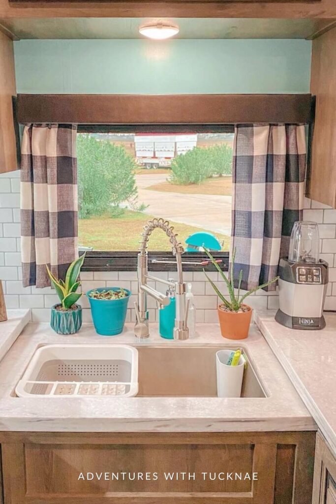 A sunny RV kitchen window dressed with checkered curtains, featuring a white sink, turquoise kitchen accessories, and a photo frame on the countertop.