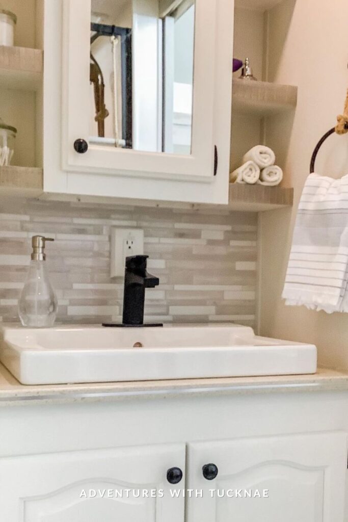 Neutral-toned RV bathroom featuring a white square sink, black faucet, light wood open shelving, mosaic backsplash, and rolled towels for spa-like organization.