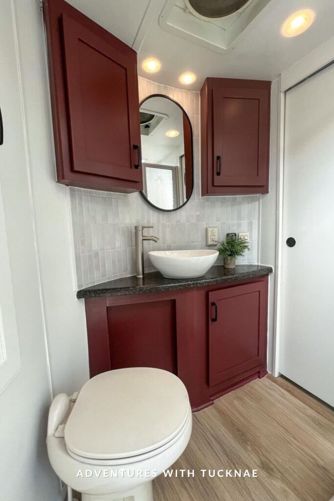 RV bathroom with bold burgundy cabinetry, granite countertop, white vessel sink, brushed nickel faucet, and a tall oval mirror between two cabinets.