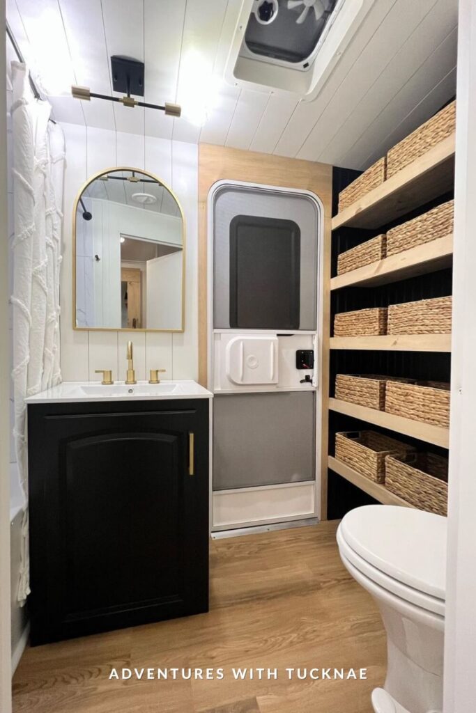 Bright RV bathroom featuring a black vanity, arched gold mirror, modern gold faucet, and custom shelving with woven baskets for added storage next to the toilet.