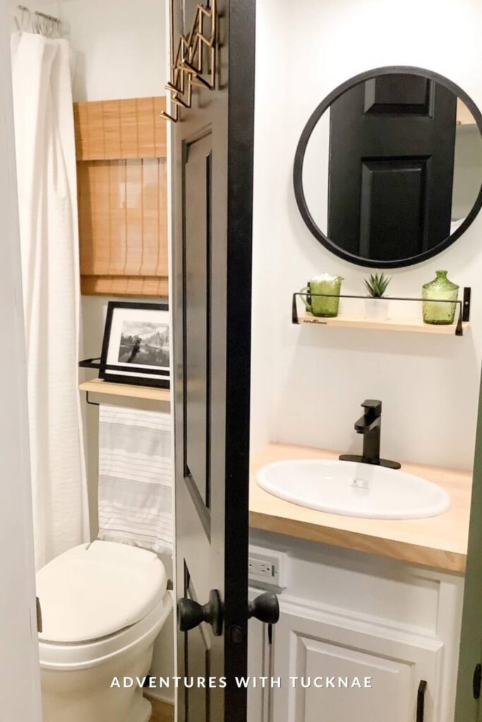 Compact RV bathroom with white cabinetry, round mirror, open wood shelves, green accents, and a black faucet on a light wood counter beside the toilet.