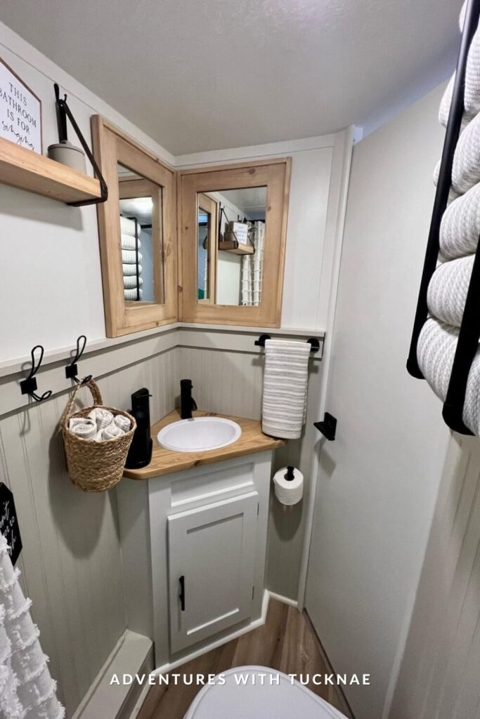 Corner vanity in an RV bathroom with a white sink, wood counter, light paneling, black fixtures, and dual mirrors in rustic wood frames above.