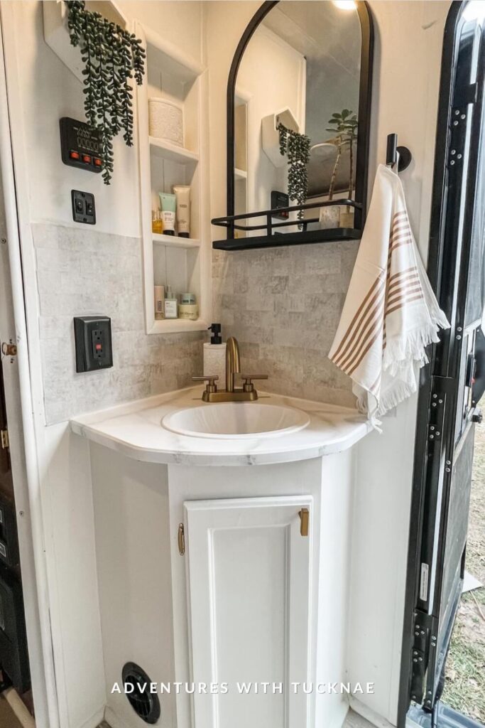Small RV bathroom with a white corner vanity, gold faucet, black-rimmed mirror, and built-in shelving filled with skincare and toiletries.
