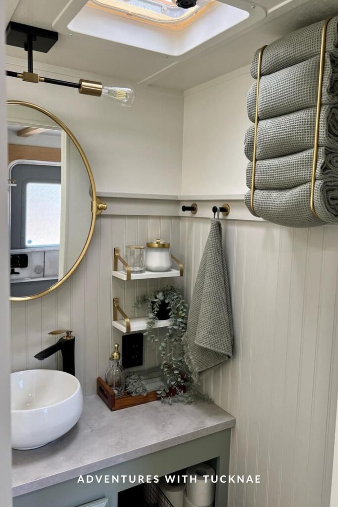 Soft, serene RV bathroom with light sage vanity, round gold mirror, black faucet, open shelving, and neatly stacked sage green towels held by gold towel bars.