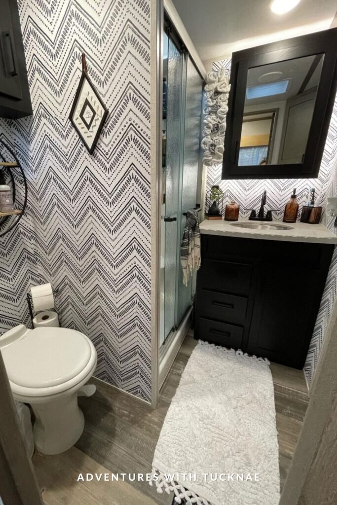 RV bathroom with bold black-and-white chevron wallpaper, black vanity, textured glass shower doors, and earthy decor featuring amber bottles and framed art.