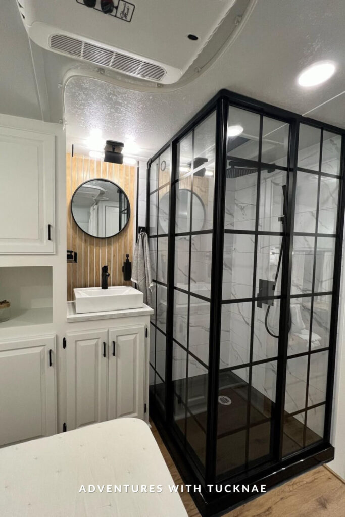 Modern RV bathroom with a black-framed glass shower, white cabinetry, circular mirror with wood accent wall, and black hardware for a sleek, updated look.