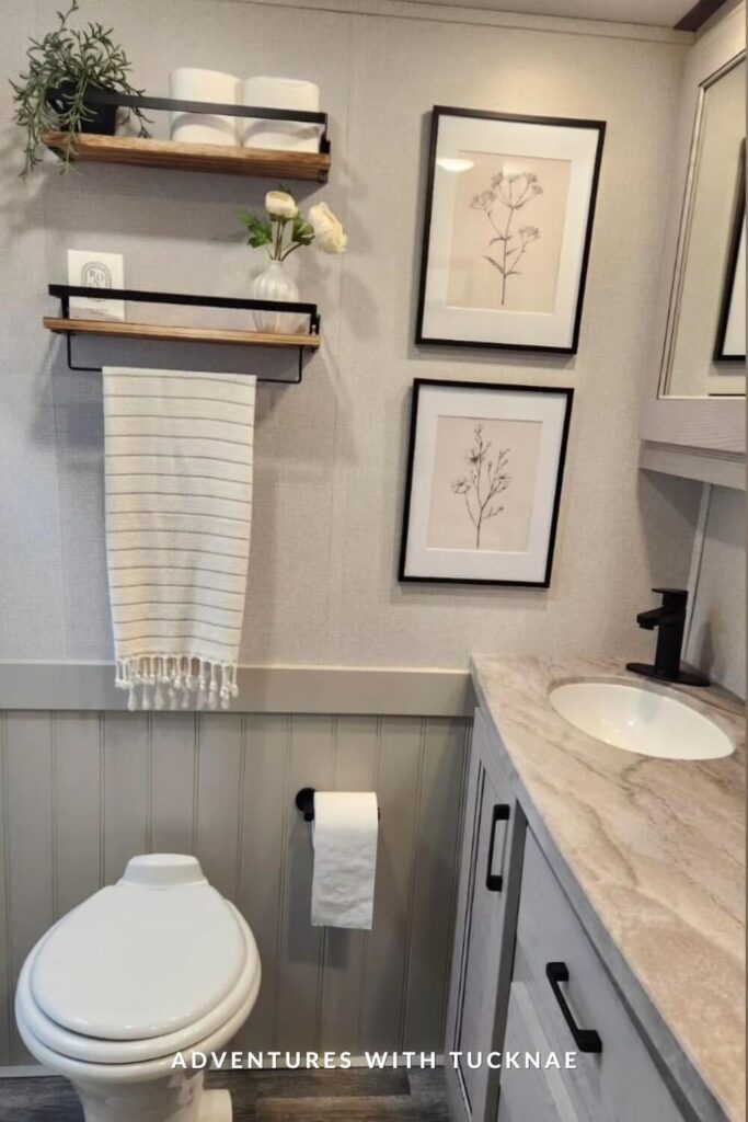 Subtle and clean RV bathroom with a light wood vanity top, black faucet, botanical wall art, floating shelves, and striped hand towel beside a white toilet.