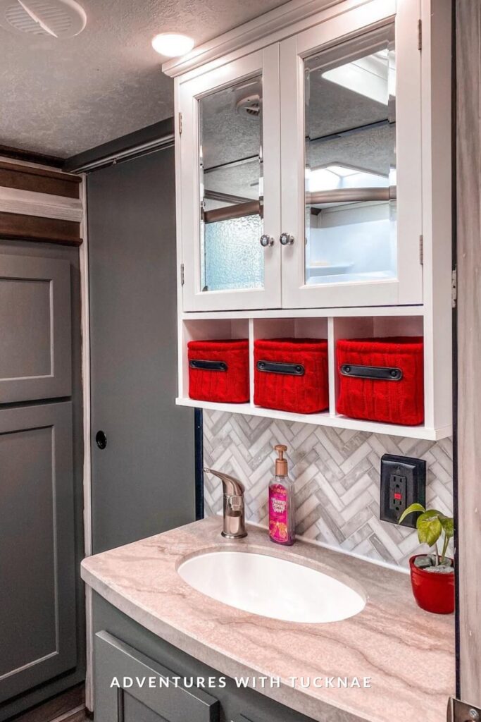 Stylish RV bathroom vanity with a marble-look countertop, brushed nickel faucet, and mirrored cabinet above, featuring bright red knit storage baskets and a herringbone tile backsplash.