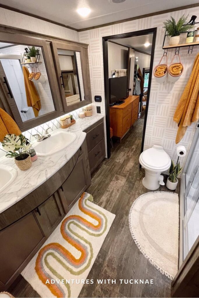 Spacious RV bathroom with dual sinks, marble-look counter, rich wood cabinets, and orange accents throughout, including rolled towels, planters, and retro-style rugs.
