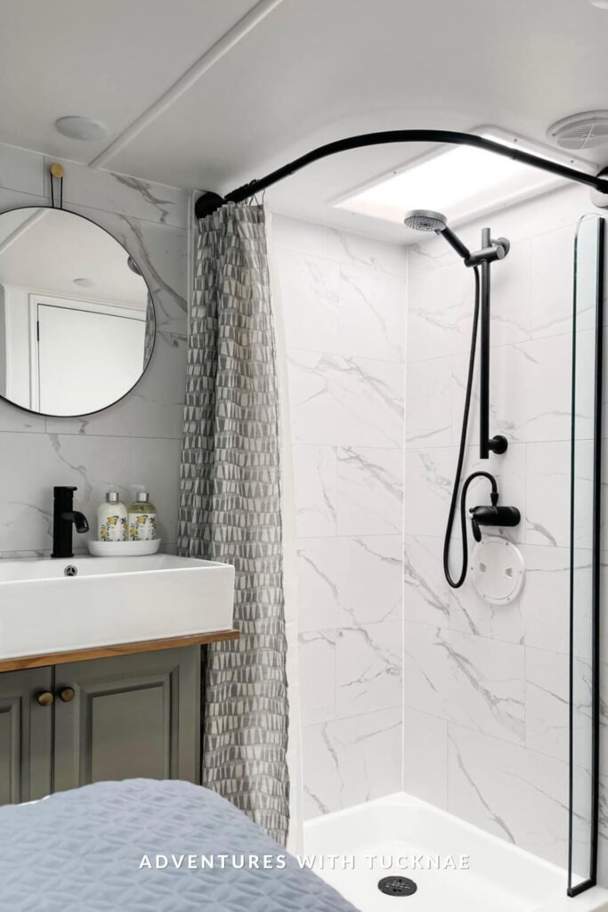 Bright RV shower space with white marble-style tiles, a curved black curtain rod, rainfall showerhead, and modern vanity with a vessel sink and round mirror.