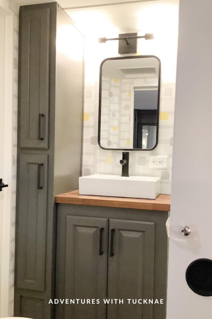 Compact RV vanity setup with a rectangular vessel sink, black faucet, matte black framed mirror, and geometric-patterned wallpaper with gray and yellow accents.