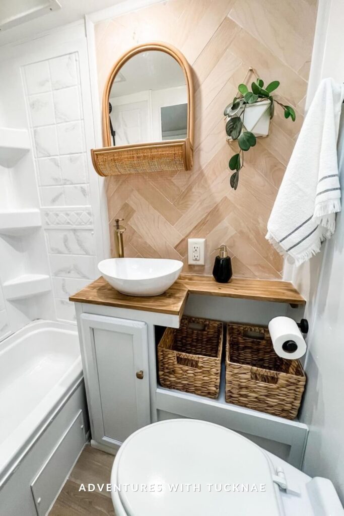 Stylish RV bath area with a herringbone wood accent wall, open wood shelving, gold faucet, and woven baskets under a wood countertop with round mirror above.