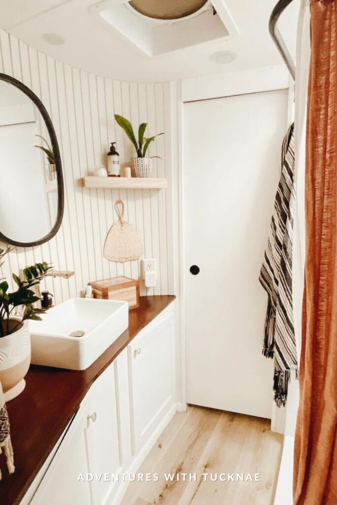 Warm-toned RV bathroom with vertical beadboard walls, wood counter, square sink, and boho accents like plants, woven wall art, and a striped towel.