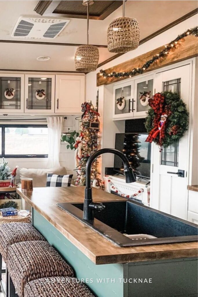 A festively decorated RV kitchen with white cabinetry, a green island with wood countertops, and rattan pendant lights. Mini wreaths and garland adorn the cabinets, and a slim Christmas tree wrapped in lights and red ribbons stands near a faux fireplace.