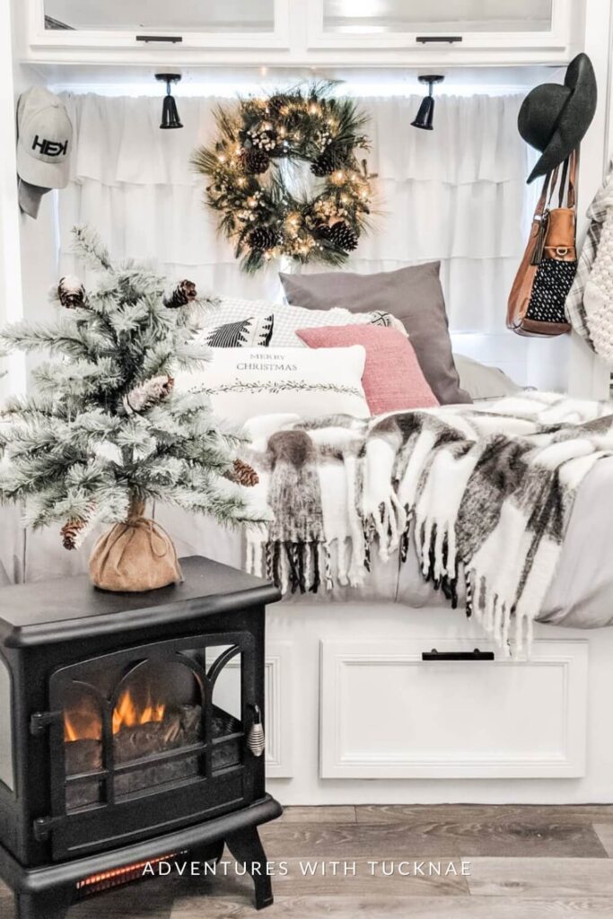 A bright, white RV nook with a fluffy throw blanket, pinecone-studded mini tree, and a wreath glowing above the bed. Neutral holiday pillows and a compact black heater add warmth and charm to the cozy space.