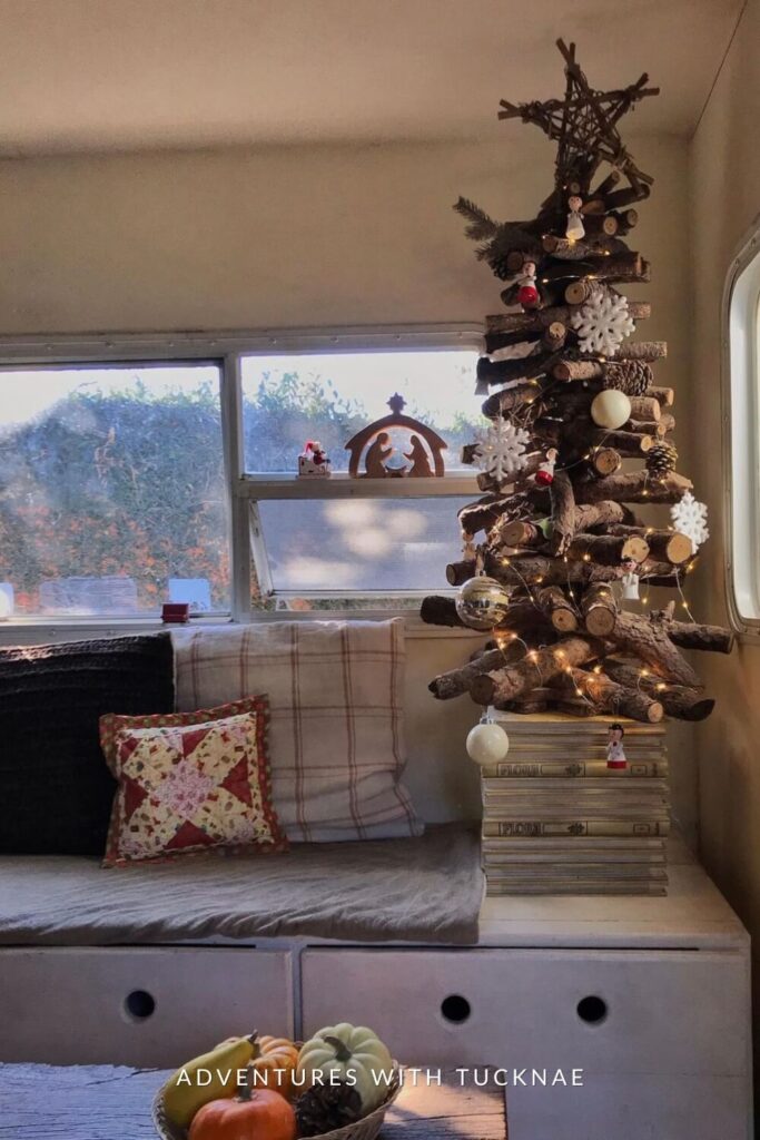 A creative, rustic-style Christmas tree made of stacked wooden branches stands in front of an RV window. Decorated with white snowflakes, miniature ornaments, and lights, the tree rests on a pile of books, adding charm to the cozy corner bench.