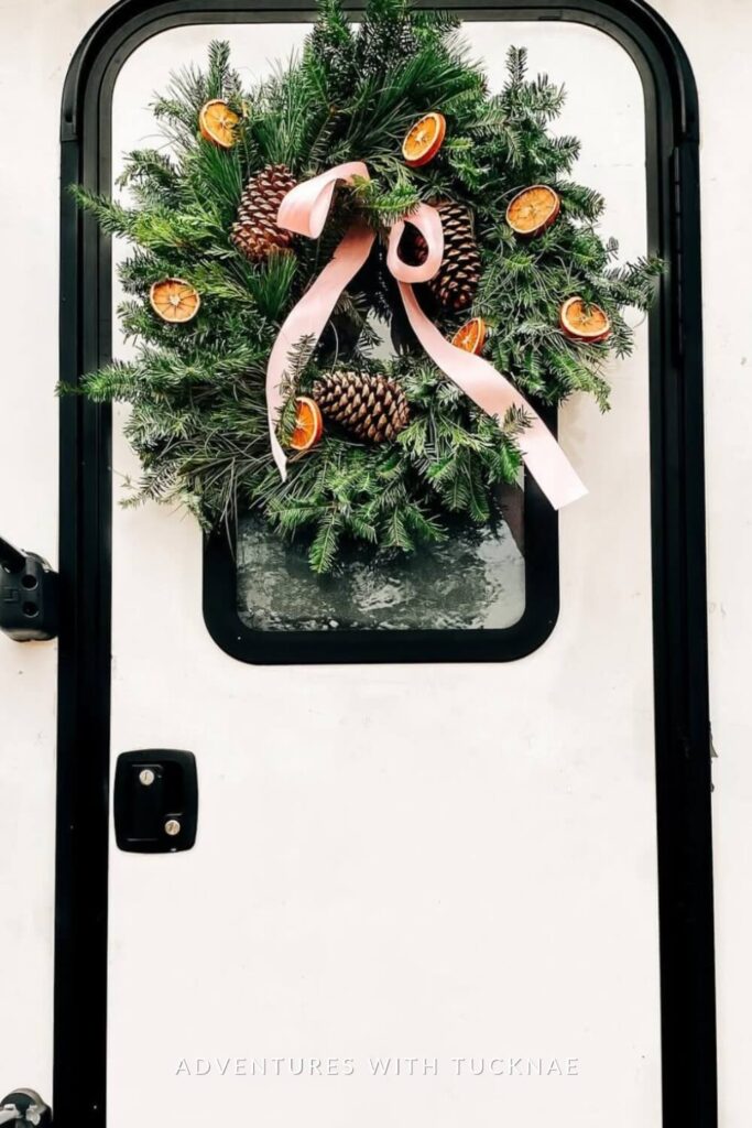 A festive green wreath with pinecones, dried orange slices, and a flowing pink ribbon is mounted on an RV door. The natural elements pop against the clean white door, bringing a handcrafted holiday feel.