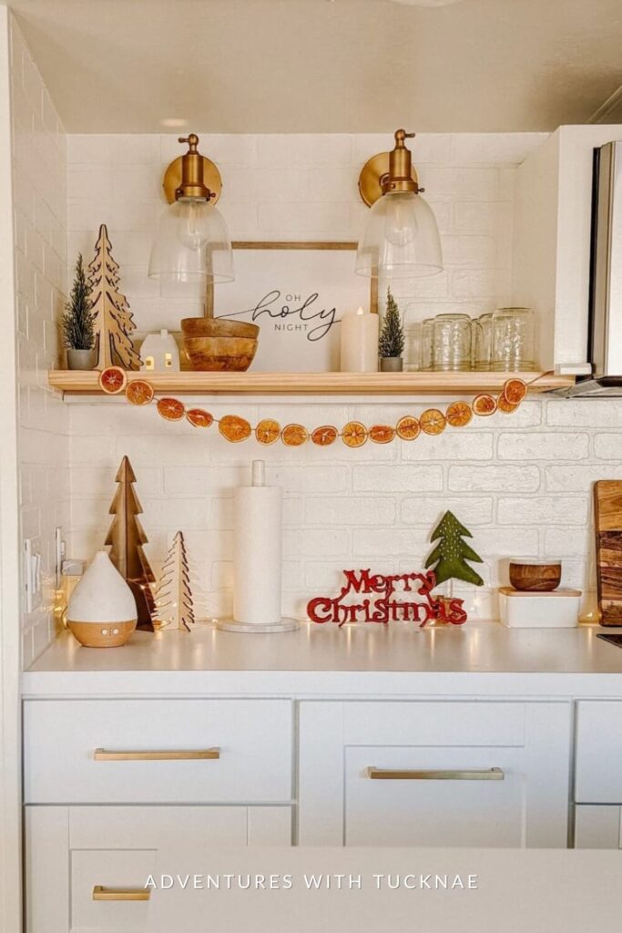 A minimalist RV kitchen shelf styled with bottle brush trees, a wood bowl, and candles, framed by a “Merry Christmas” sign and orange slice garland. The white shiplap and warm lighting give it a clean, Scandinavian holiday vibe.