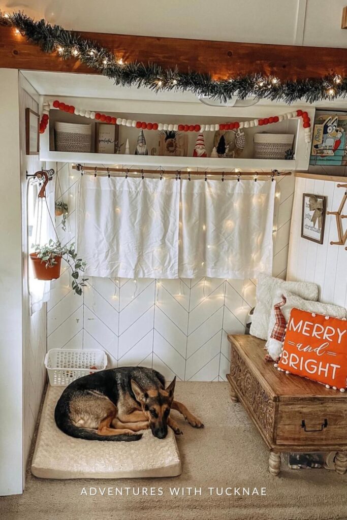 A festive nook inside an RV with a garland-strung shelf, red and white felt ball garland, and mini gnomes decorating the upper cubby. Below, a German Shepherd lies on a soft dog bed beside a bench with a “Merry and Bright” pillow and string lights.