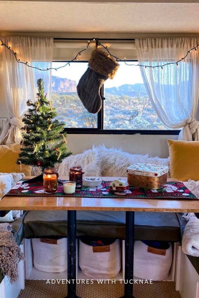 A dining area inside an RV decorated with a mini Christmas tree, candles, and a red holiday table runner. A large stocking hangs from fairy lights strung above the window, which opens to a scenic mountain view.