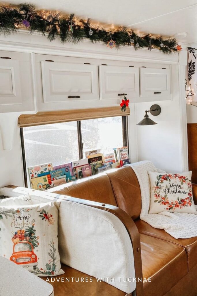 A light-filled RV living space featuring a brown leather couch with “Merry Christmas” pillows, a kids’ bookshelf below the window, and garland with twinkle lights running across the white cabinetry above.