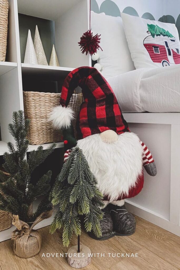 A whimsical Christmas gnome with a red and black plaid hat and white beard sits next to miniature pine trees. Behind him are woven baskets and cone-shaped tree decor on a cubby shelf, with a camper-themed holiday pillow in the background.