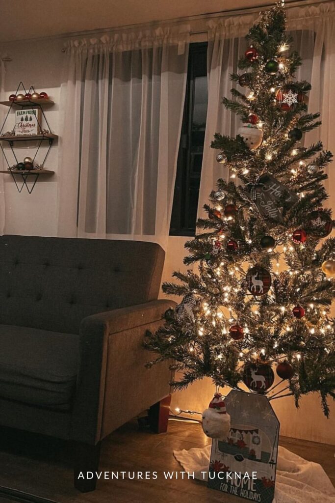 A warmly lit RV interior with a Christmas tree glowing in the corner, decked out with red, white, and black ornaments and a “Ho Ho Jolly” sign. Behind it, sheer curtains frame a dark gray sofa and two holiday-themed wall shelves.