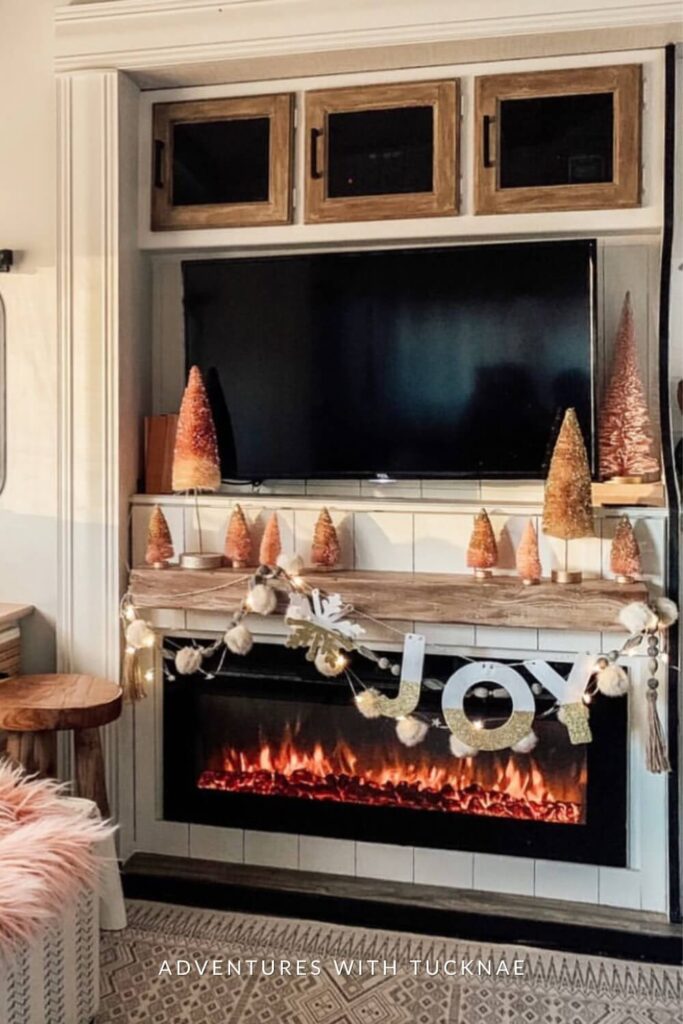 A festive RV fireplace setup with a cozy electric flame beneath a wooden mantel decorated with bottle brush trees in shades of pink and gold. A “JOY” garland with glitter letters, pom-poms, and snowflake cutouts hangs across the front, creating a warm and cheerful holiday vibe.