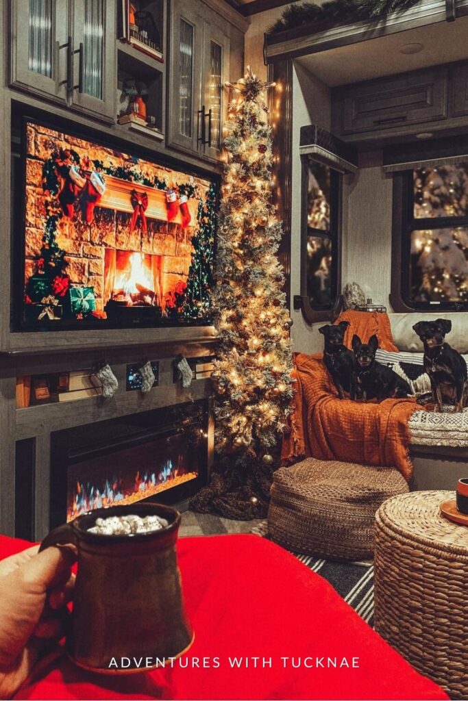 A holiday-decorated RV living space with a faux fireplace below a large TV displaying a virtual yule log. A lit, frosted Christmas tree stands in the corner, and three small black dogs sit on a blanket-draped bench while someone holds a mug of hot cocoa in the foreground.