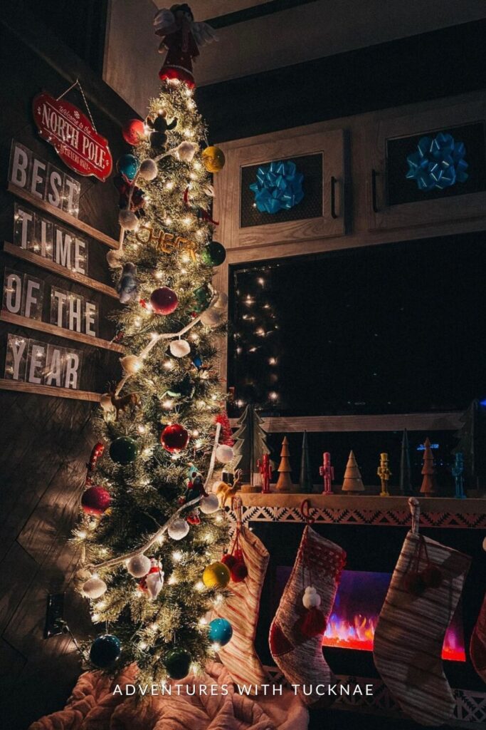 A tall, narrow Christmas tree decorated with multicolored ornaments and a topper angel stands in a cozy RV corner. Stockings hang beneath a shelf of wooden trees, while wall signs read "Welcome to the North Pole" and "Best Time of the Year."