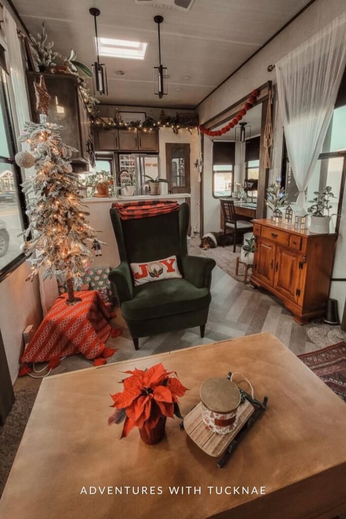 A rustic-modern RV setup with a green armchair featuring a “JOY” pillow, and a snowy mini tree set on a red tablecloth. The space is dressed with garland, soft lighting, and holiday details like wrapped presents and poinsettias.