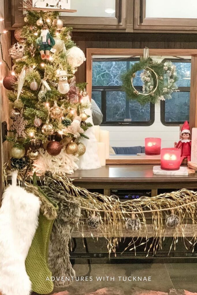 A glam and playful Christmas vignette with a mini tree full of unique ornaments, including owls and glittery baubles. Nearby are red candles, a green wreath in the window, and stockings in various textures and colors draped with gold fringe garland.