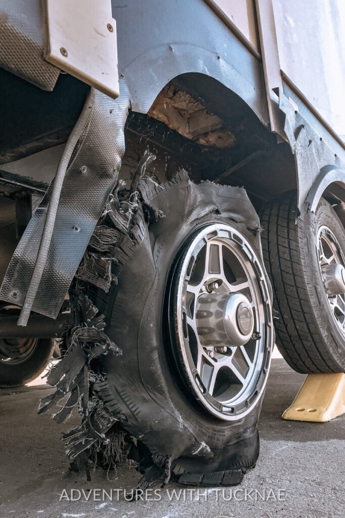 Close-up of a shredded RV tire after a blowout, with torn rubber and fabric hanging in jagged strips and visible damage to the wheel well. The dramatic scene underscores the importance of RV tire maintenance and safety checks.