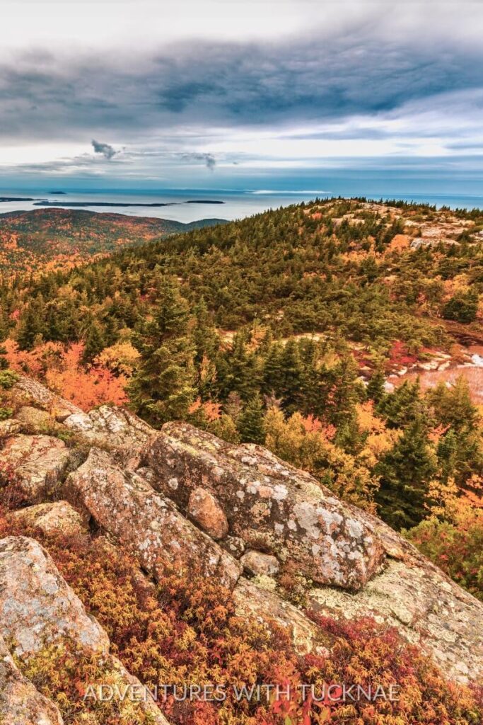 View from a rocky overlook in Acadia National Park with a sweeping vista of fall-colored forests and distant ocean islands under a dramatic cloudy sky. A quintessential example of scenic beauty for fall RV destinations in New England.