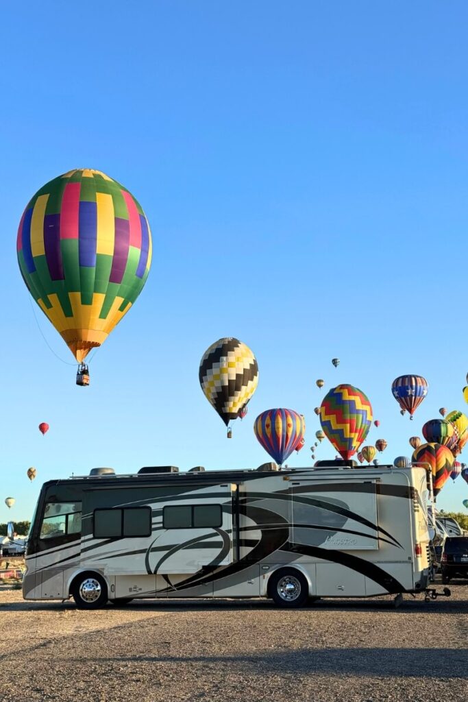 Hot air balloons in vibrant colors float above a luxury RV during the Albuquerque International Balloon Fiesta, set against a clear blue sky. A festive and unique highlight for fall RV destinations in the Southwest.