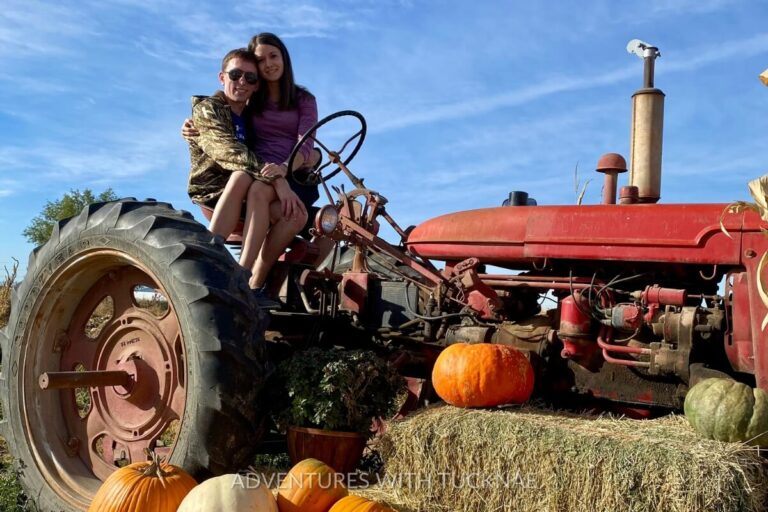Tucker and Janae sit together on an old red tractor surrounded by hay bales and a variety of pumpkins in a festive autumn farm setting.